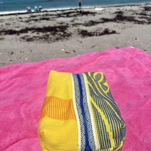 May include: A rectangular fabric pouch featuring a vibrant yellow and orange woven texture on one side and a blue and white patterned design on the other, separated by a central blue zipper closure that runs vertically down the middle of the item.