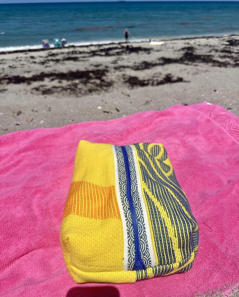 May include: A rectangular fabric pouch featuring a vibrant yellow and orange woven texture on one side and a blue and white patterned design on the other, separated by a central blue zipper closure that runs vertically down the middle of the item.