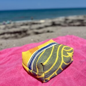 May include: A rectangular zippered pouch featuring a vibrant yellow and orange fabric with a dark blue leaf-like pattern. The pouch has a distinct blue zipper closure and a decorative white and blue patterned trim along the top edge of the bag.