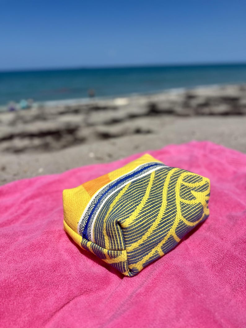 May include: A rectangular zippered pouch featuring a vibrant yellow and orange fabric with a dark blue leaf-like pattern. The pouch has a distinct blue zipper closure and a decorative white and blue patterned trim along the top edge of the bag.