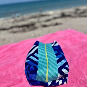 May include: A rectangular zippered pouch featuring a blue and white geometric pattern on the sides and a central vertical panel of teal and light blue wavy stripes, accented by a bright yellow zipper running down the center of the pouch.