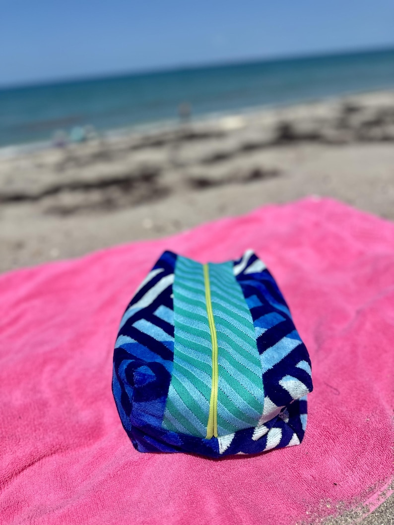 May include: A rectangular zippered pouch featuring a blue and white geometric pattern on the sides and a central vertical panel of teal and light blue wavy stripes, accented by a bright yellow zipper running down the center of the pouch.