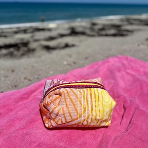 May include: A small, rectangular fabric pouch featuring a vibrant pattern of yellow, orange, and pink lines. The pouch has a bright yellow zipper closure accented by a dark purple trim with a repeating geometric pattern along the top edge.