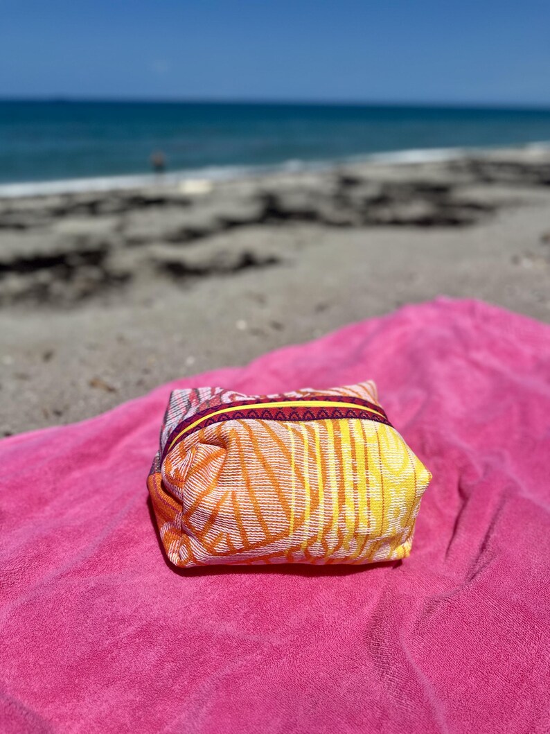 May include: A small, rectangular fabric pouch featuring a vibrant pattern of yellow, orange, and pink lines. The pouch has a bright yellow zipper closure accented by a dark purple trim with a repeating geometric pattern along the top edge.
