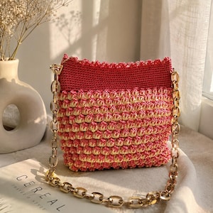 May include: A crochet handbag featuring a textured bobble stitch pattern in alternating horizontal stripes of pink and cream yarn. The bag has a solid pink top border with a scalloped edge and is equipped with a thick, polished gold-tone chain strap.