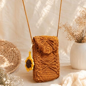 Pode incluir: Uma bolsa transversal de crochê na cor amarelo mostarda com um padrão geométrico texturizado na aba frontal e no corpo. A bolsa inclui uma alça longa combinando e um pequeno pingente removível em formato de girassol com pétalas amarelas.
