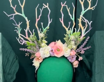 May include: A pastel rainbow colored floral headband with white, pink, and purple flowers. The headband is made of branches and has a green background.