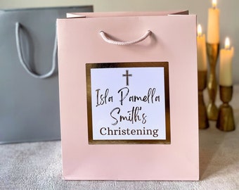Può includere: Una borsa regalo rosa con una croce dorata e il testo "Isla Pamela Smith's Christening" su un'etichetta bianca.