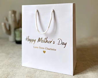 Puede incluir: Bolsa de regalo blanca con letras doradas que dicen "Feliz Día de la Madre" y "Amor de Charlotte" con un pequeño corazón dorado.