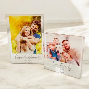 May include: Two clear acrylic photo blocks with personalised text and photos. The first block features a couple embracing in a sunny outdoor setting with the text "Katie & Daniel 12.7.2022". The second block shows a family with a dog and the text "Our Family 2024".