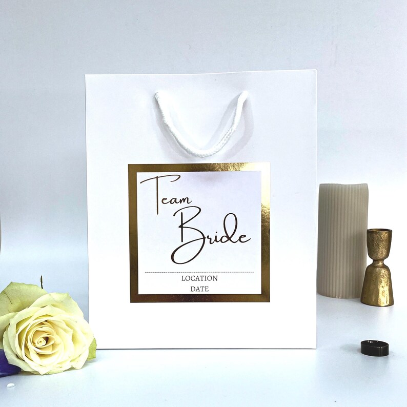Luxury Personalised Team Bride Gift Bag and white sticker with gold foil writing and sticker. Writing says team bride with the option for adding a name and a date or location. Perfect for hen party favours and hen party gifts for your Team Bride.