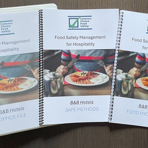 May include: Three white binders with spiral binding. Each binder has a photo of a person eating a breakfast meal. The text on the binders reads "Food Safety Management for Hospitality" and "Caterers Choice Food Safety". The binders are labeled "Office File", "Safe Methods", and "Food Hygiene Diary".