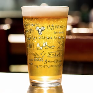May include: A pint glass filled with golden beer and a foamy head. The glass is adorned with handwritten scientific equations and symbols. The glass is sitting on a reflective surface.