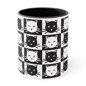 May include: A white ceramic mug with a black interior. The exterior features a repeating pattern of black and white cat faces in a checkerboard design. The cat faces alternate between open-eyed and crossed-out eyes.