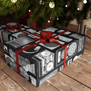 May include: A black gift box wrapped in paper featuring a pattern of vintage audio equipment, such as turntables, cassette players, and speakers. The box is tied with a red ribbon.