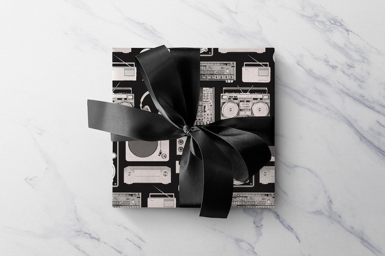 May include: A black gift box with a black ribbon tied in a bow. The box is wrapped in wrapping paper with a pattern of vintage audio equipment, including turntables, cassette players, and boomboxes.