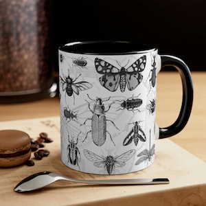 Peut inclure: Une tasse en céramique blanche avec un bord et une poignée noirs. La tasse présente une illustration en noir et blanc de divers insectes et papillons, dont un papillon de nuit, un scarabée et une libellule.