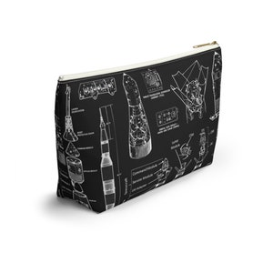 May include: Black cosmetic bag featuring a white diagram of a rocket and spacecraft components. The bag has a gold zip and a white interior. The diagram includes labels such as "Command Module" and "Lunar Module".