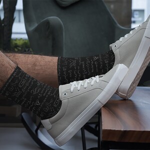 K&ouml;nnte beinhalten: Hellgraue Sneaker mit wei&szlig;en Sohlen und Schn&uuml;rsenkeln, kombiniert mit schwarzen Socken mit wei&szlig;en wissenschaftlichen Diagrammen. Die Schuhe liegen auf einer Holzoberfl&auml;che. Die Sneaker sind ein l&auml;ssiger Stil.