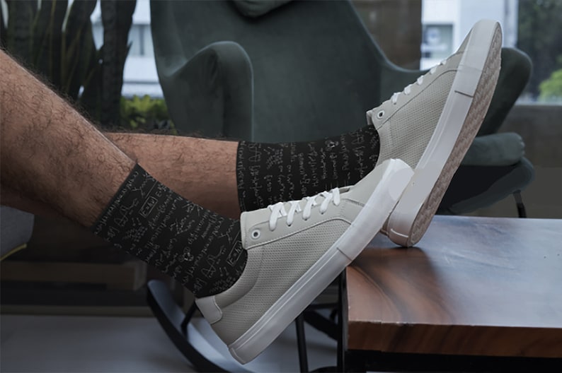 K&ouml;nnte beinhalten: Hellgraue Sneaker mit wei&szlig;en Sohlen und Schn&uuml;rsenkeln, kombiniert mit schwarzen Socken mit wei&szlig;en wissenschaftlichen Diagrammen. Die Schuhe liegen auf einer Holzoberfl&auml;che. Die Sneaker sind ein l&auml;ssiger Stil.