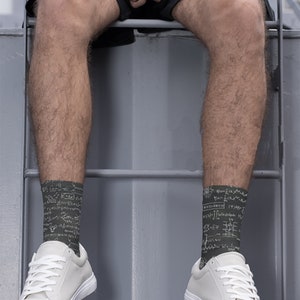 K&ouml;nnte beinhalten: Eine Person tr&auml;gt dunkelgr&uuml;ne Socken mit wei&szlig;en mathematischen Gleichungen und wei&szlig;en Turnschuhen. Die Socken sind der Mittelpunkt, wobei die Gleichungen die gesamte Socke bedecken. Die Turnschuhe sind wei&szlig; mit wei&szlig;en Schn&uuml;rsenkeln.