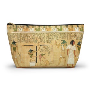Puede incluir: Una bolsa de cosméticos beige con cierre de cremallera presenta una imagen de una escena del antiguo Egipto. La escena representa una ceremonia de pesaje del corazón con Anubis, el dios con cabeza de chacal, que pesa el corazón de una persona fallecida contra una pluma. La escena está rodeada de jeroglíficos.