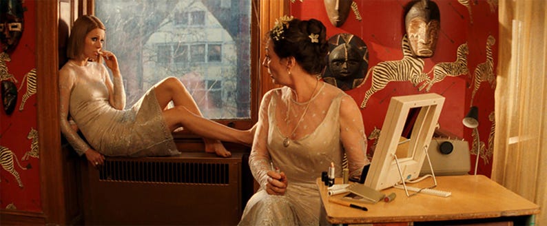 May include: Two women in a room with red and white zebra patterned wallpaper. One woman is sitting on a windowsill wearing a silver sequined dress. The other woman is sitting at a table with a mirror and makeup supplies. She is wearing a white dress with a beaded necklace.