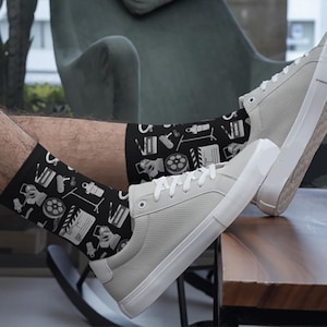 May include: A pair of black socks with a white film-themed pattern. The socks are being worn by a person wearing white trainers.
