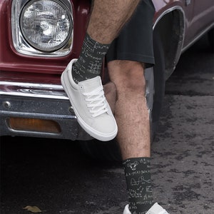 K&ouml;nnte beinhalten: Schwarze Socken mit wei&szlig;en mathematischen Gleichungen und Symbolen, getragen mit wei&szlig;en Turnschuhen. Die Socken sind an den Unterschenkeln sichtbar, die Schuhe ruhen auf einer dunklen Oberfl&auml;che. Im Hintergrund ein Oldtimer.