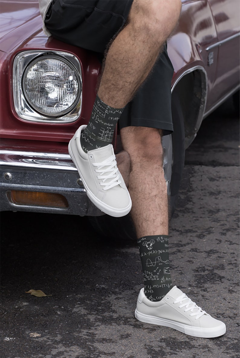 K&ouml;nnte beinhalten: Schwarze Socken mit wei&szlig;en mathematischen Gleichungen und Symbolen, getragen mit wei&szlig;en Turnschuhen. Die Socken sind an den Unterschenkeln sichtbar, die Schuhe ruhen auf einer dunklen Oberfl&auml;che. Im Hintergrund ein Oldtimer.