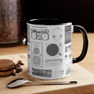 May include: A black and white mug with a black handle. The mug features a white design of various music equipment, including a boombox, speakers, headphones, and a record player.