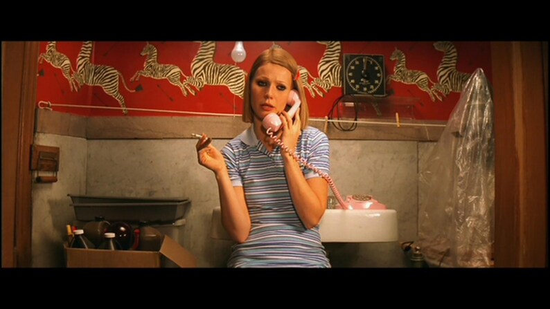 May include: A woman with blonde hair is sitting in a bathroom and talking on a pink rotary phone. She is wearing a striped blue and white shirt and a blue and white striped skirt. The bathroom has a red and white zebra print wallpaper.