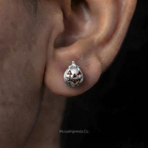 May include: A silver-toned stud earring shaped like a carved pumpkin, featuring small, dark orange stones set into the triangular eyes, nose, and jagged mouth. The text Hussainjewels.co is printed in small white letters at the bottom of the image.
