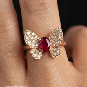May include: A gold ring featuring a butterfly-shaped design with a central oval-cut deep red ruby gemstone surrounded by small clear pavé-set diamonds on the wings. The text Hussainjewels.Co is visible in the bottom left corner of the image.