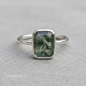 Puede incluir: Un anillo de plata con una piedra rectangular de ágata musgosa. La piedra verde translúcida tiene inclusiones de color verde oscuro, engastada en un bisel de plata. La banda del anillo es de plata pulida.