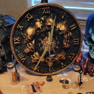 Könnte beinhalten: Eine schwarze und goldene Uhr mit Totenkopfmotiv. Das Zifferblatt der Uhr hat römische Ziffern und ein Zahnrad-Design in der Mitte. Die Uhr steht auf einem Holzsockel mit Totenkopfdesign.
