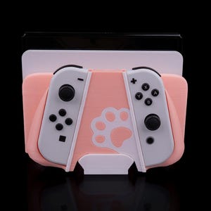 Può includere: Un supporto rosa e bianco per console Switch con un design a zampa. Il supporto contiene la console e due controller, grigi e bianchi. Il supporto è in plastica e ha uno sfondo nero.