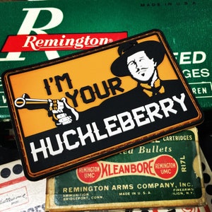 May include: A black and orange patch with a cartoon illustration of a man wearing a hat and holding a gun. The text on the patch reads "I'm Your Huckleberry".