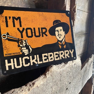 May include: A black and orange metal sign with the text "I'm Your Huckleberry" and a cartoon image of a man with a gun. The sign has a distressed look and the text "PATRIOT PATCH CO." is visible in the bottom left corner.