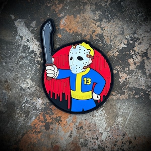 May include: A black and red circular patch with a cartoon character wearing a blue jumpsuit, a yellow vest, and a hockey mask. The character is holding a large knife and the number "13" is on the vest. The text "PATRIOT PATCH CO." is on the bottom of the patch.
