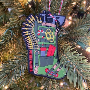 May include: A green Christmas stocking ornament with a variety of tactical gear, including a candy cane, a knife, a brass knuckle, and a magazine of bullets. The stocking is decorated with the text "PATRIOT PATCH CO."