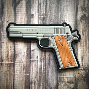 May include: A gray and orange patch in the shape of a handgun. The patch has the words "PATRIOT 45 AUTO" printed on the side. The grip is orange with a textured pattern, and the patch is outlined in black.