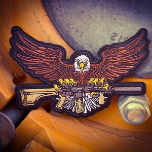 May include: A brown and black embroidered patch of an eagle holding a rifle with its talons. The eagle has a yellow beak and yellow talons. The rifle is black with a brown stock. The patch is on a yellow and brown background.