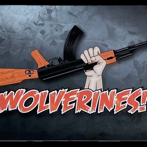 May include: A metal sign with a cartoon image of a hand holding a rifle with a wooden stock. The text "WOLVERINES!" is printed in red and white letters.
