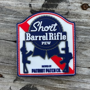 May include: A rectangular patch with a red, white, and blue border. The patch features the words "Short Barrel Rifle" and "PEW" in white text, along with two blue rifle silhouettes. The text "BREWED BY PATRIOT PATCH CO." is also included.