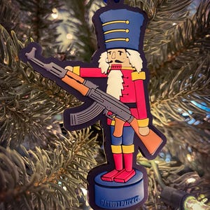 May include: A Christmas ornament shaped like a nutcracker holding a rifle. The nutcracker is wearing a red and blue uniform with a white beard and mustache. The ornament is made of rubber and has a blue ribbon attached to the top. The ornament has the text "PATRIOT PATCH CO." on the base.