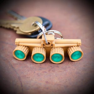 May include: A tan-colored keychain featuring a design with four cylindrical elements with teal-colored centers. The keychain is attached to a silver keyring, with keys visible in the background. The design is reminiscent of a scientific instrument.