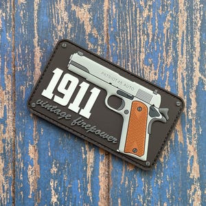 May include: A rectangular patch featuring a silver and orange handgun with the text "1911 vintage firepower" and "PATRIOT 45 AUTO". The patch is brown with silver rivets and is set against a blue and brown wood grain background.