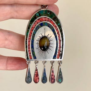 以下が含まれることがあります： 太陽光線デザインに囲まれた、中央の楕円形のタイガーアイストーンが付いたシルバーペンダント。ペンダントは、赤、青、緑のカラフルなモザイクアーチデザインが特徴です。下部からは5つのしずく型のダングルがぶら下がっています。