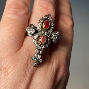 May include: An ornate silver ring featuring two oval red gemstones and numerous small clear gemstones. The ring has a floral design with a central cluster and delicate leaf-like accents. The ring is worn on a finger.